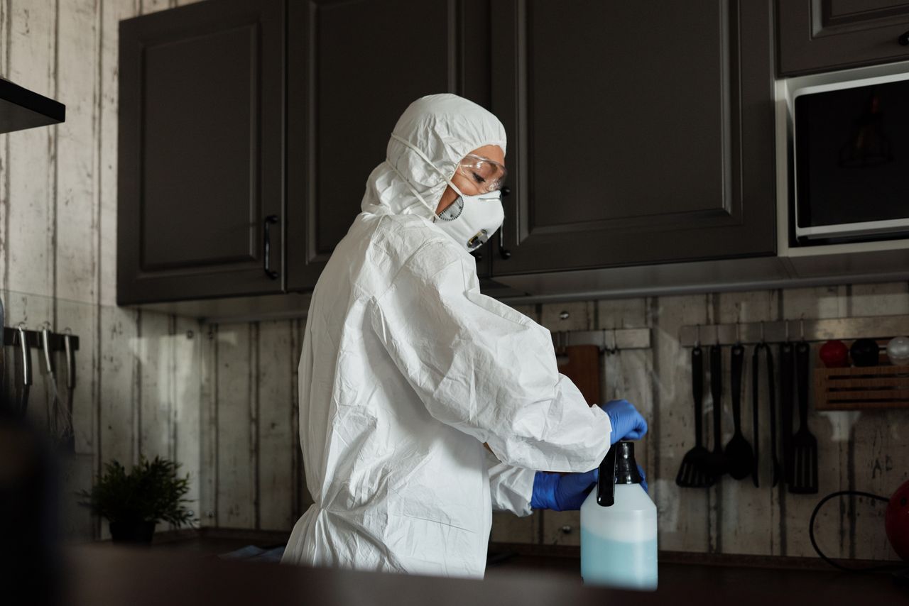 Services-03 Healthcare worker in PPE uses disinfectant spray in a kitchen setting during COVID-19 pandemic.